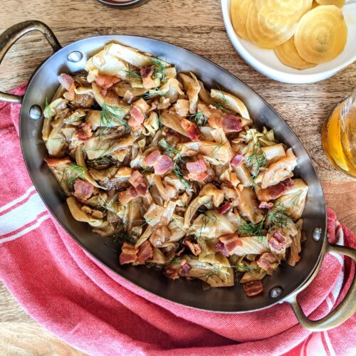 An oval bronze baking dish full of apple cider vinegar braised cabbage and fennel. Thick pieces of applewood smoked bacon are sprinkled throughout. Garnished with fennel fronds.
