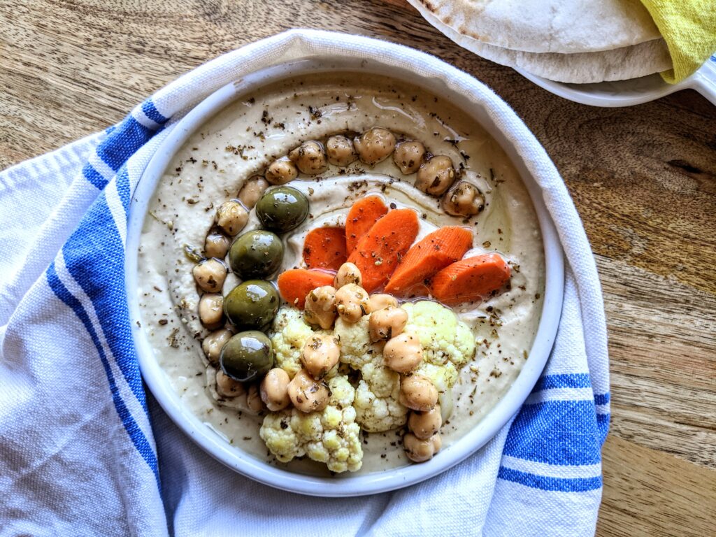 A shallow bowl of creamy hummus, topped with whole chickpeas, green olives, and homemade carrot and cauliflower pickles.