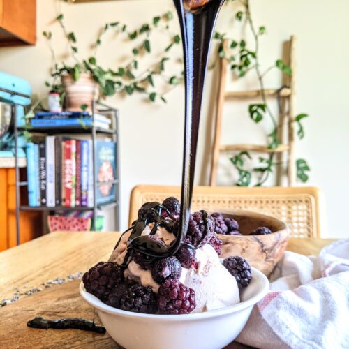 Thick and rich balsamic glaze dripping from a spoon onto a cup of ice-cream and frozen berries.