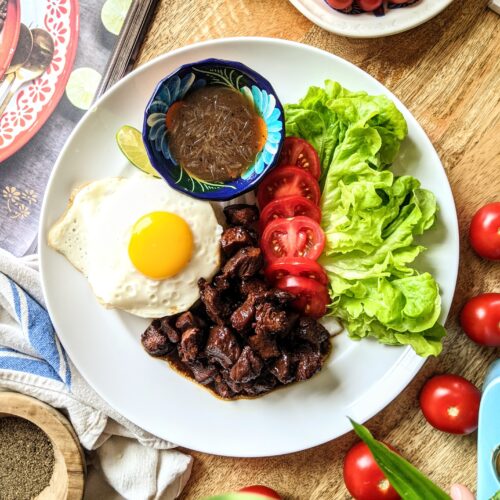 A plate with marinated pieces of beef lok lak, a small bowl of lime pepper sauce, pieces of leafy green lettuce, sliced tomatoes and onion, and a lightly fried egg sitting on top of Jasmin rice.