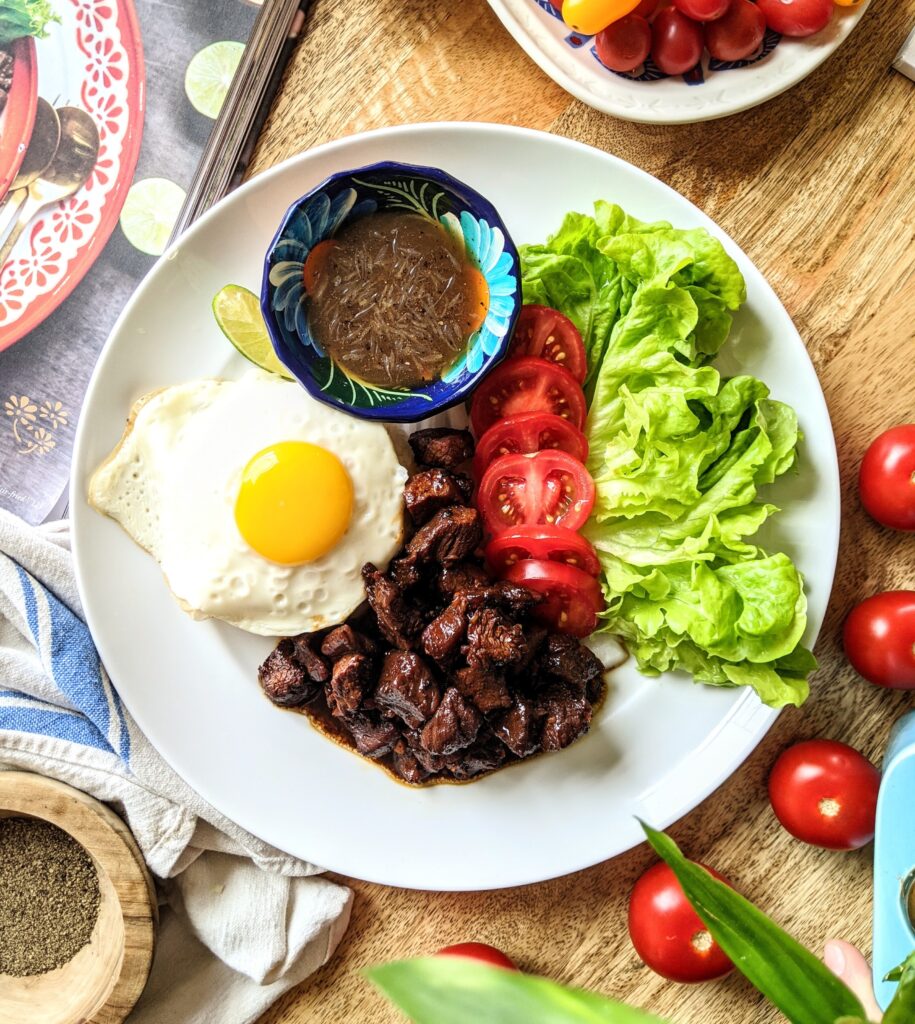 A plate with marinated pieces of beef lok lak, a small bowl of lime pepper sauce, pieces of leafy green lettuce, sliced tomatoes and onion, and a lightly fried egg sitting on top of Jasmin rice.