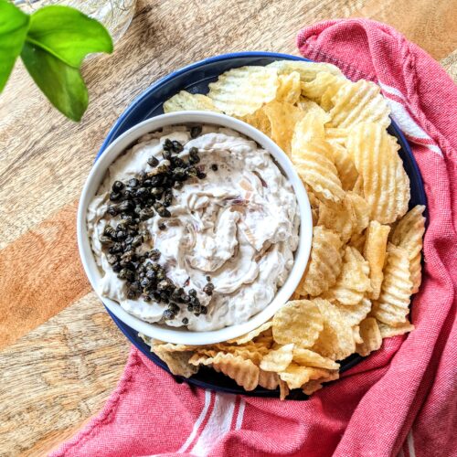 A bowl of rich buttermilk french onion dip topped with crispy fried capers, served alongside thick ridged potato chips.