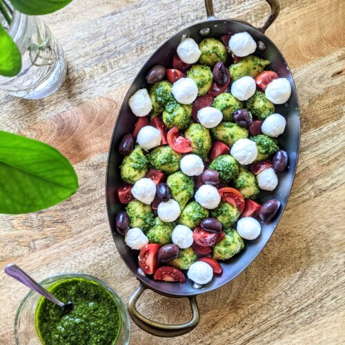 An oval baking dish full of soft gnocchi, small mozzarella balls, tomatoes, Kalamata olives, and fresh basil pesto; ready for the oven!