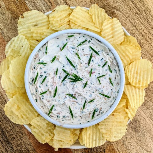 A bowl of caramelized shallot and chive dip, garnished with sliced chives and a sprig of dill. Served in the center of a plate and surrounded by wavy original potato chips.