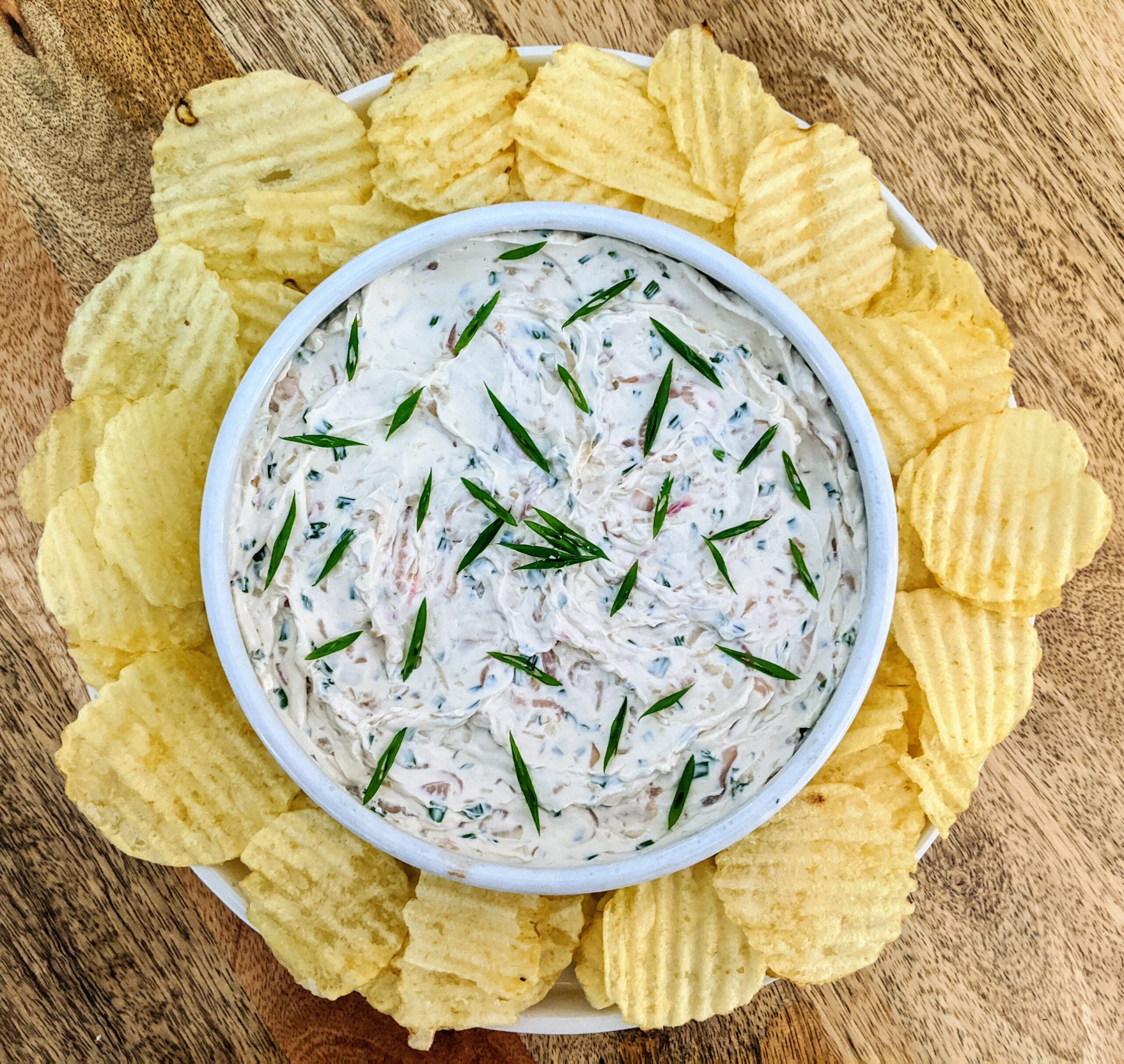 A bowl of caramelized shallot and chive dip, garnished with sliced chives and a sprig of dill. Served in the center of a plate and surrounded by wavy original potato chips.