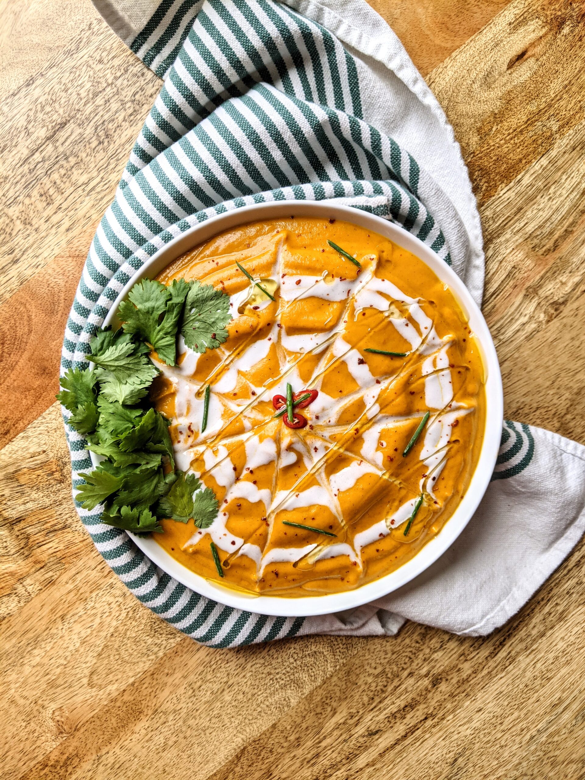 A bowl of vibrant orange carrot coconut curry soup, with an intricate spider web-like design of coconut cream, garnished with fresh cilantro, chives, and sliced Thai chili.