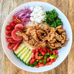 A colorful Greek chicken gyro bowl, featuring diced bell peppers, sliced cucumber, quartered small tomatoes, pickled radishes, red onion slivers, crumbled feta, and chopped fresh dill.