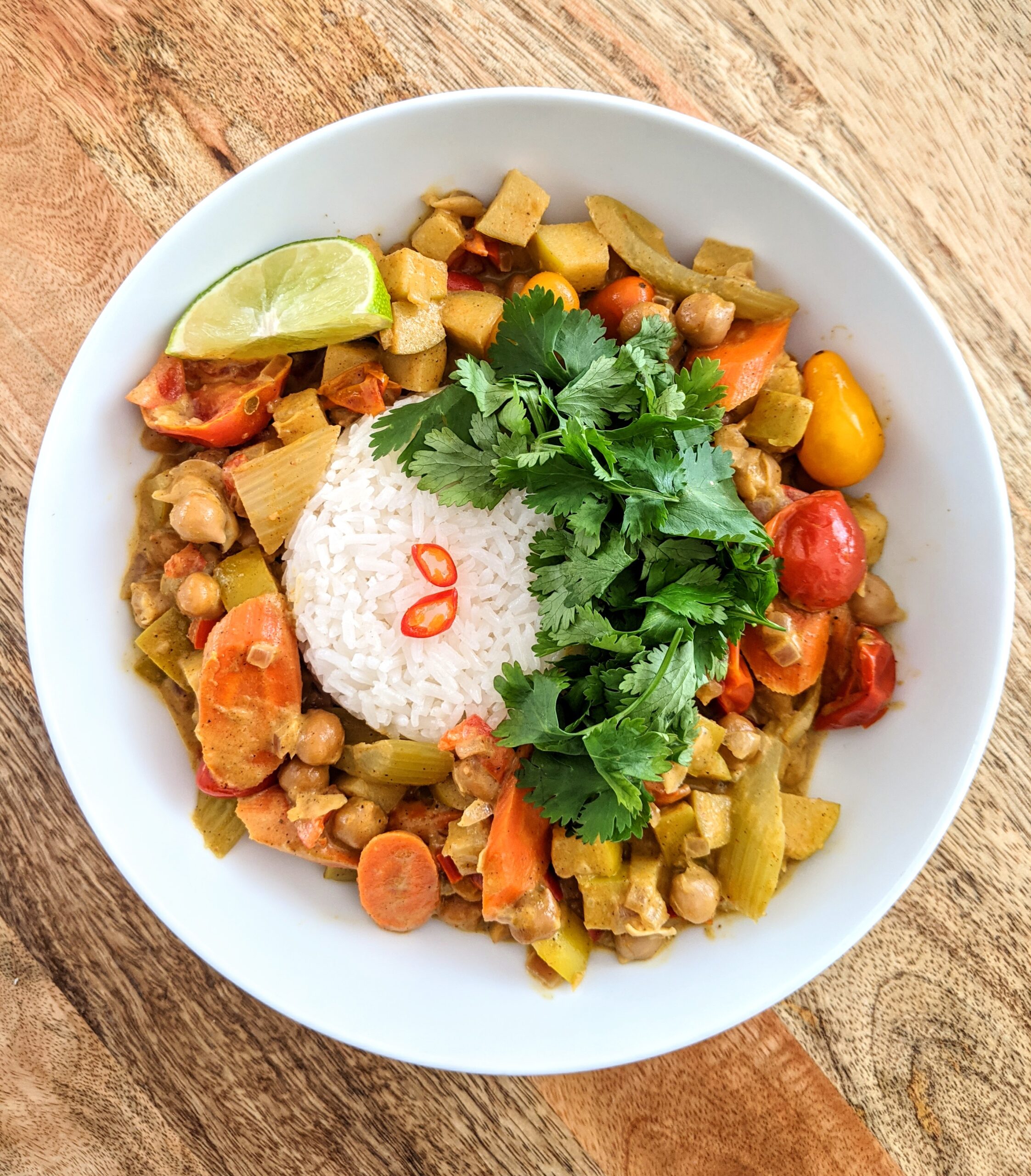 A bowl of chickpea and apple coconut curry, plated around an anchor of Jasmine rice. Topped with loads of fresh cilantro and sliced Thai chili. Served with a lime wedge.