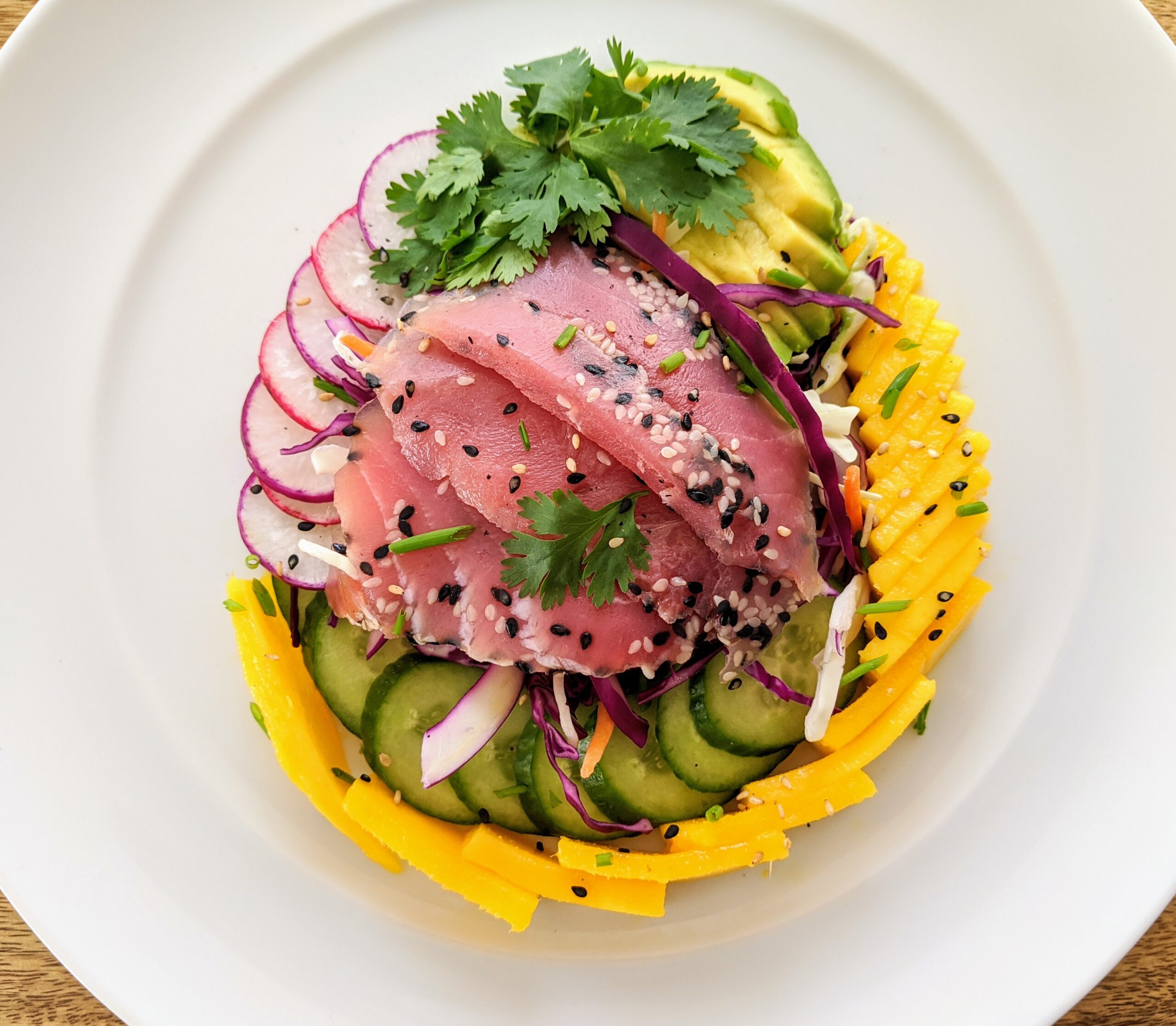 A tear dropped shaped salad with Ahi Tuna in the center, surrounded by yellow mango, purple and pink radishes, cucumber, avocado, and cabbage slaw tossed in a Citrus and Sesame dressing. Garnished with fresh cilantro, toasted sesame seeds and more dressing on the side.