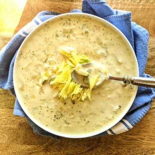 A bowl of comforting Cream of Celery Soup. A large spoonful showcasing pieces of celery, dill, celery leaves, and a thick & creamy chicken broth.