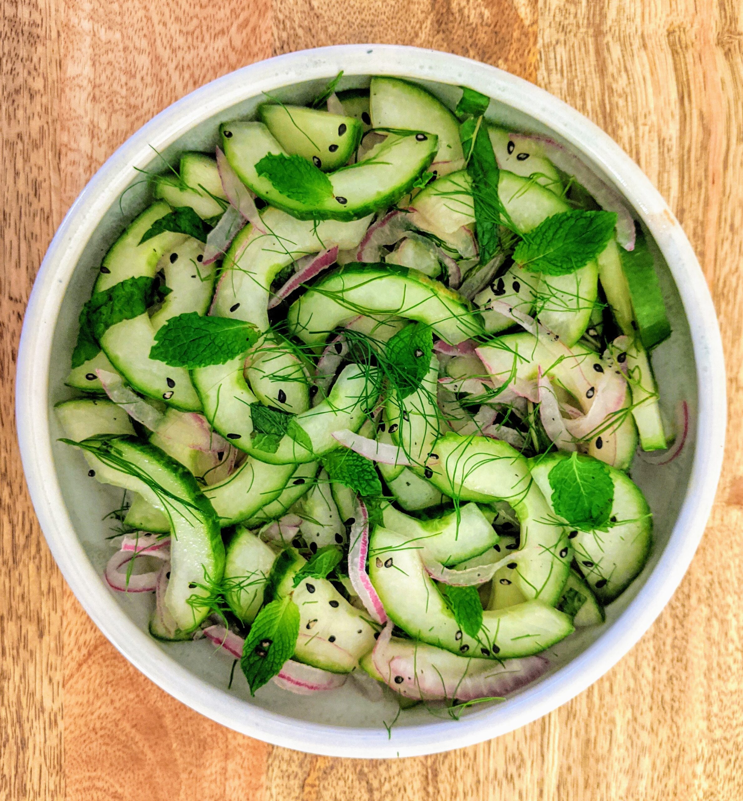 A shallow bowl full of sliced English cucumber halves, thinly sliced red onion, toasted black sesame seeds, and a trio of fresh herbs (cilantro, dill, and mint).
