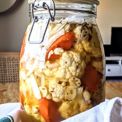 A jar of thickly sliced carrots, small cauliflower florets, thinly slice white onion, floating in a sea of pickle brine and a variety of spices.