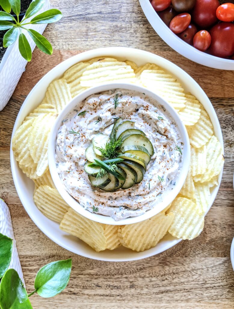 A bowl of thick dill pickle and chive dip; garnished with sliced dill pickle rounds, sliced chives, torn dill, and a sprinkling of Old Bay seasoning.. Served with wavy plain potato chips.