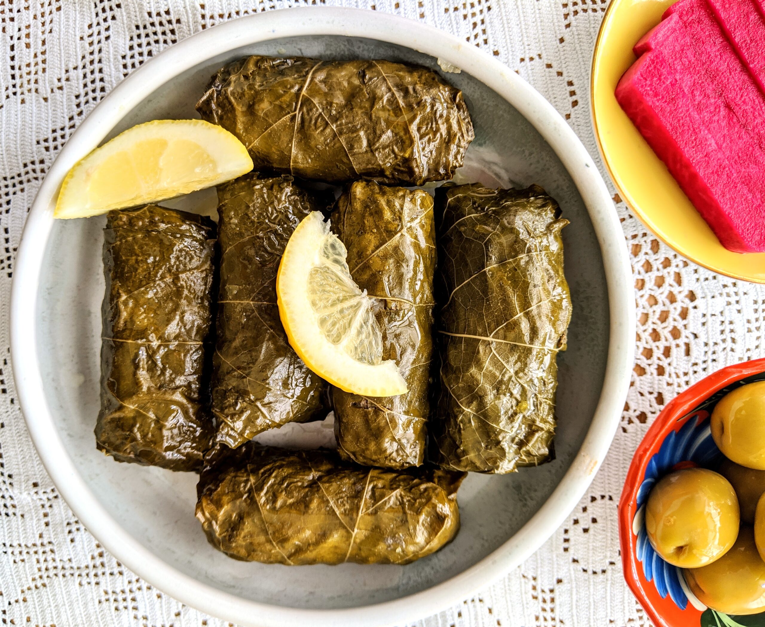 A plate full of lamb stuffed grape leaves, served with lemon wedges, bright pink pickled turnips, and large green olives.