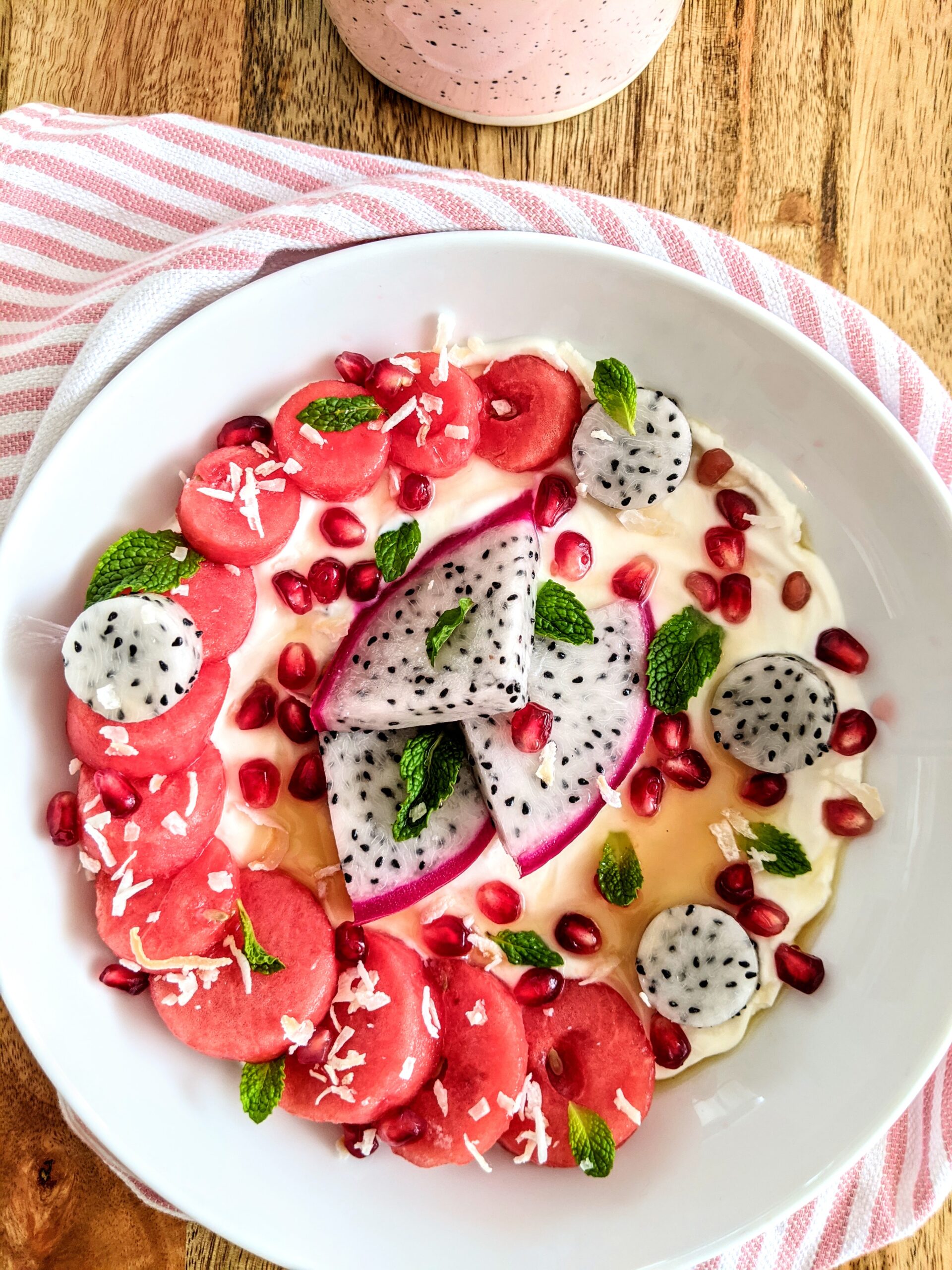 A vibrant and colorful yogurt bowl full of dragonfruit, watermelon and pomegranate. Garnished with toasted coconut flakes, honey, and mint leaves.