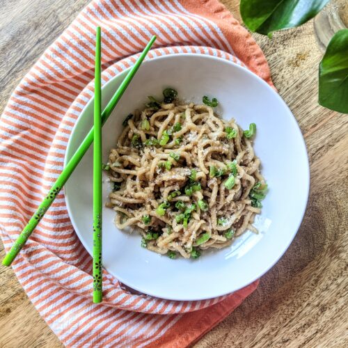 A bowl of garlic noodles garnished with sliced scallions. A set of bright green chopsticks rest on the bowl and a vibrant orange and white tea-towel wraps loosely around.