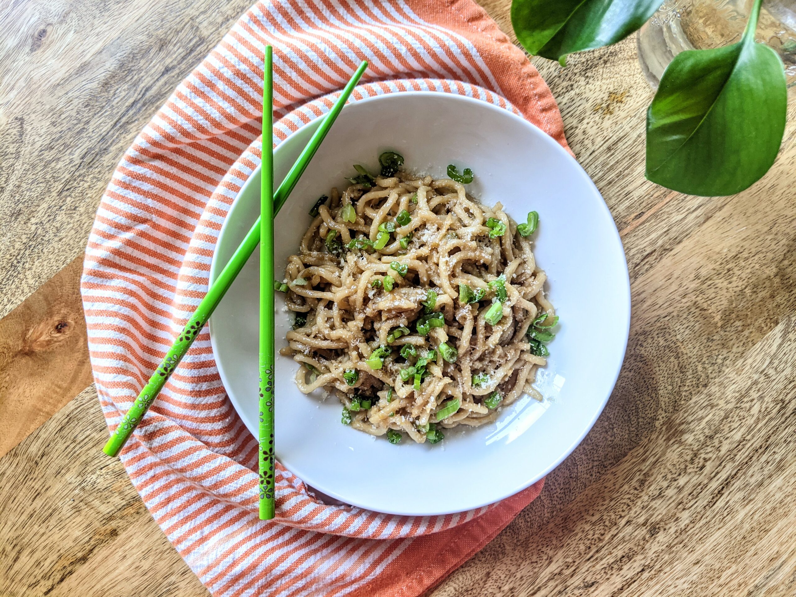 A bowl of garlic noodles garnished with sliced scallions. A set of bright green chopsticks rest on the bowl and a vibrant orange and white tea-towel wraps loosely around.