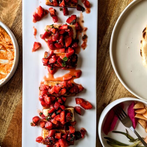 Six pieces of salty grilled Halloumi cheese, topped with sweet and spicy strawberry salsa. This dish is plated on a long white platter, surrounded by other dips and a variety of homemade pickles.