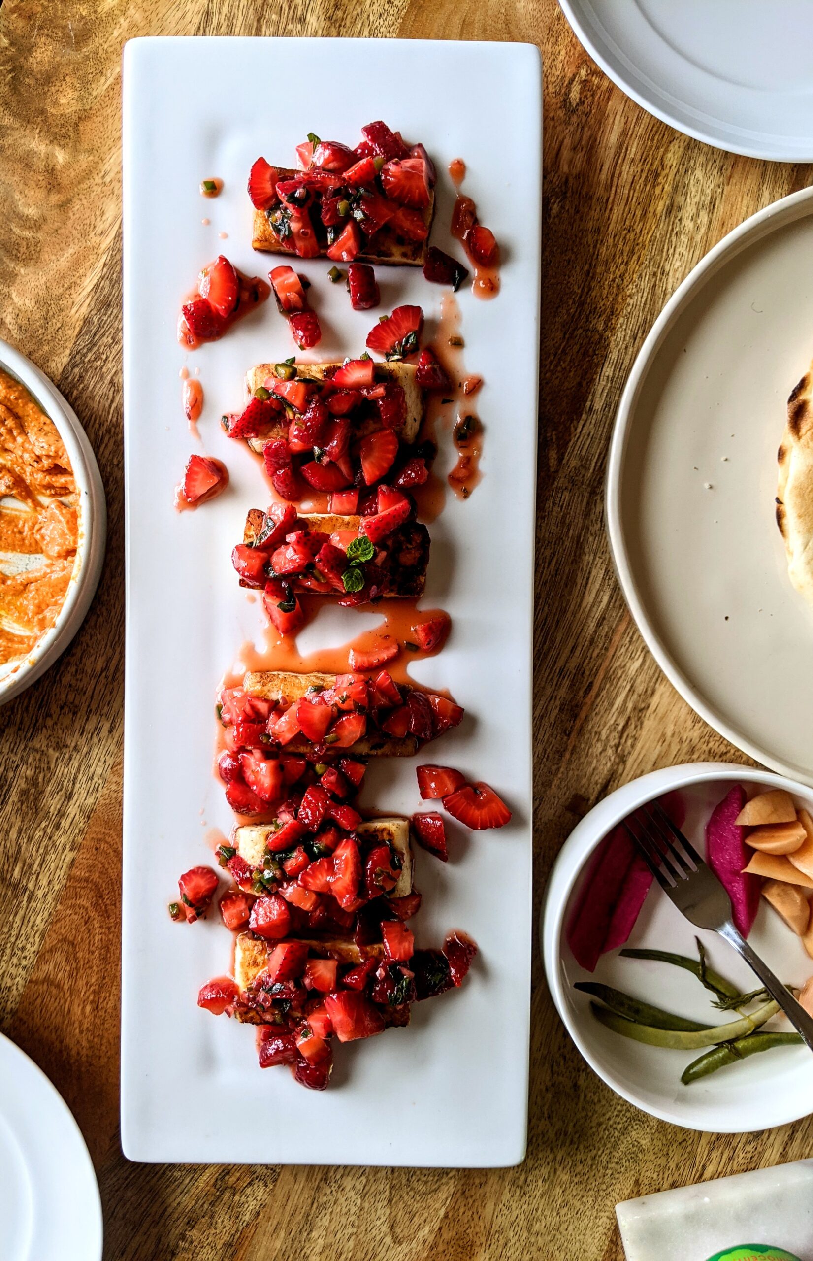 Six pieces of salty grilled Halloumi cheese, topped with sweet and spicy strawberry salsa. This dish is plated on a long white platter, surrounded by other dips and a variety of homemade pickles.
