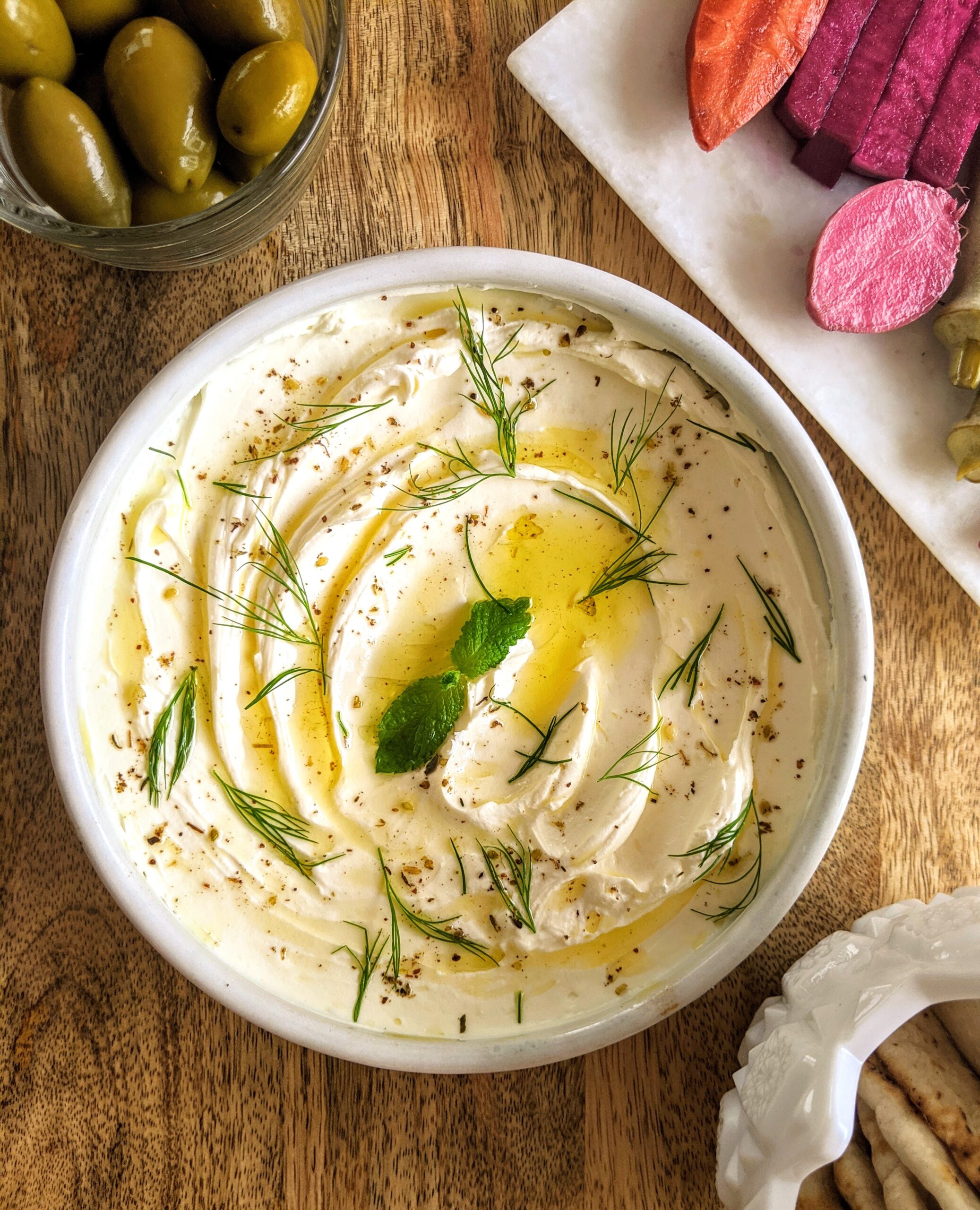 A shallow bowl of homemade labneh swooshed and swirled with a spoon to add dimension. Garnished with a drizzle of extra-virgin olive oil, mint leaves, torn dill, and a light sprinkle of za'atar. Served alongside a variety of homemade pickled vegetables, warm pita bread, and Cerignola olives.