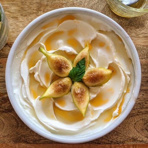 Creamy and tangy homemade labneh swooshes and swirled onto a shallow rimmed plate, extra-virgin olive oil and hot honey pool between each crevice, just waiting to be scooped up. Quartered Mission figs float on top of the yogurt cheese. Garnished with a single basil leaf.