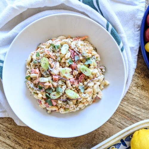 A bowl of vegetable packed macaroni salad; garnished with sliced scallions, micro greens, and poppyseeds.