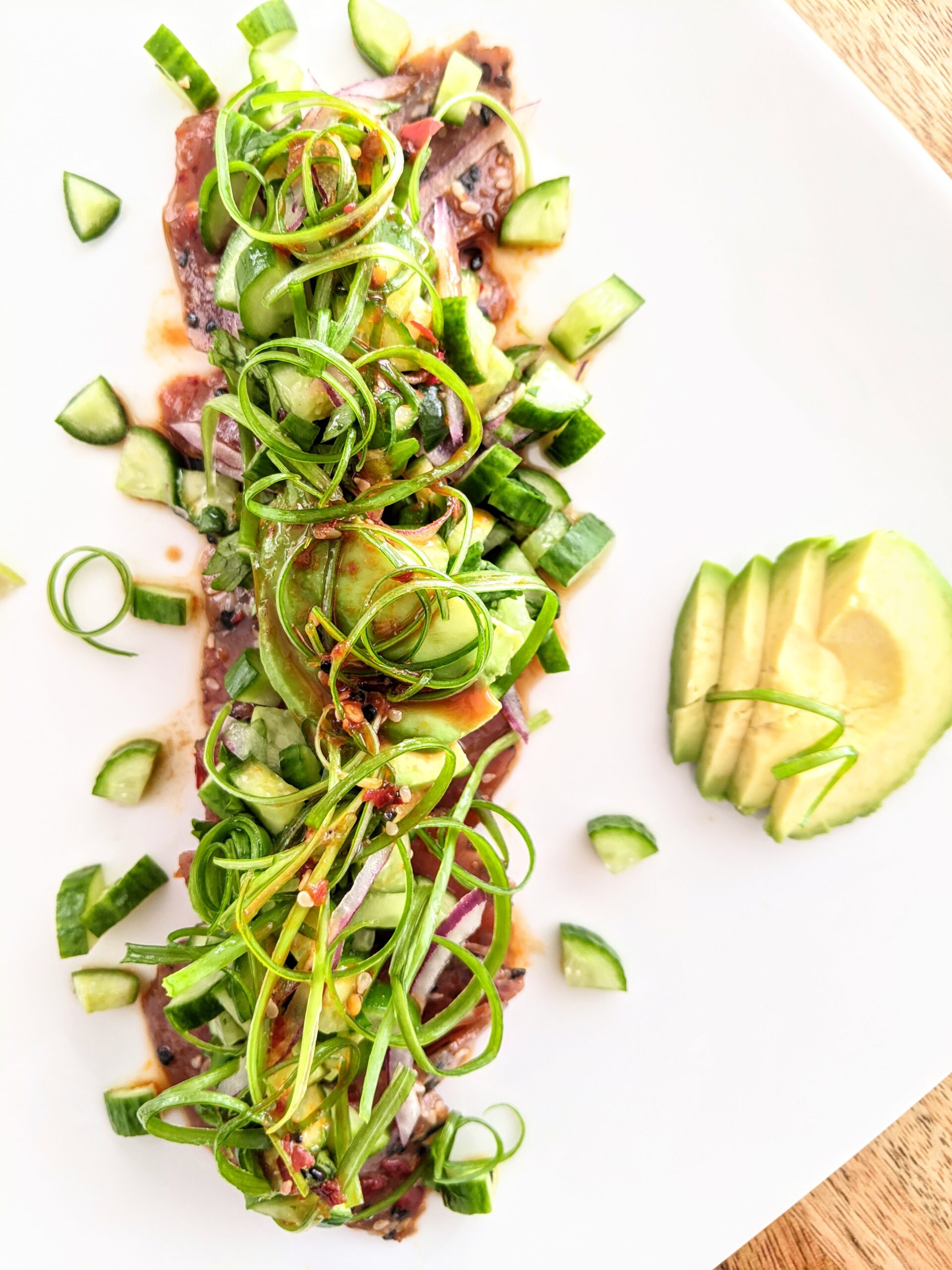 Marinated spicy ahi tuna tataki, topped and surrounded by spring onion curls, avocado slices, and diced cucumber salad.