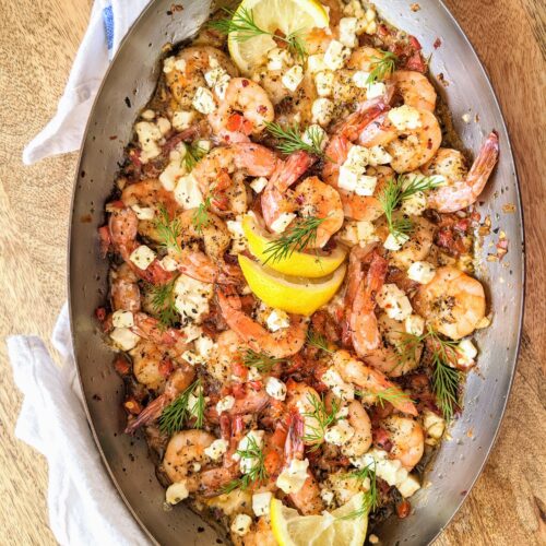An oval copper baking dish filled with juicy shrimp, wedges of lemon, finely diced red pepper, crumbled feta, and loads of fresh dill & oregano. The pan juices are just begging to be enjoyed with warm crusty bread!