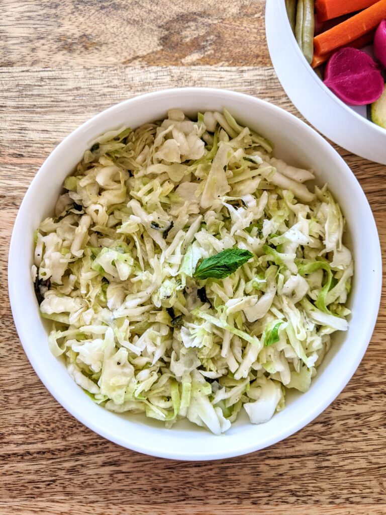 A bowl of shredded cabbage tossed in a light sauce made of garlic, extra-virgin olive oil, lemon juice, and dried fresh mint. Pictured alongside a variety of homemade pickled vegetables.