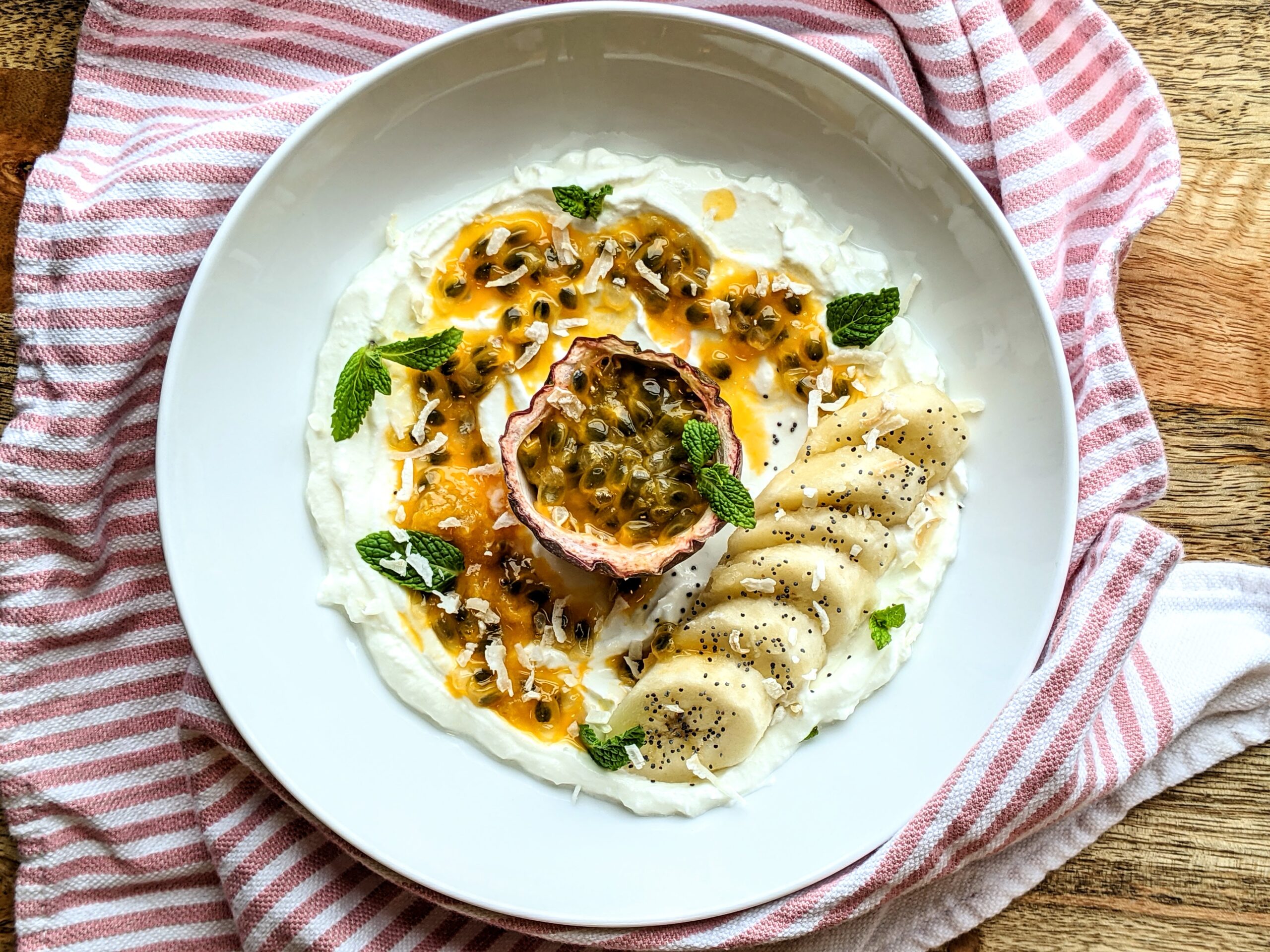 A hallow bowl of thick Greek yogurt topped with vibrant tangerine colored passionfruit pulp, sliced banana, half a pomegranate, chia seeds, and fresh mint leaves.