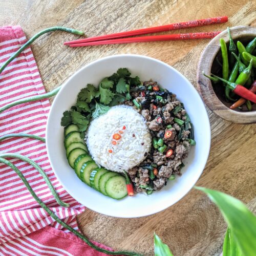 A bowl of spicy Thai basil chicken, steamed Jasmine rice, sliced cucumbers, and fresh cilantro; garnished with sliced Thai chili and fermented black garlic. Long beans and a small bowl of Thai chilies are also pictured.