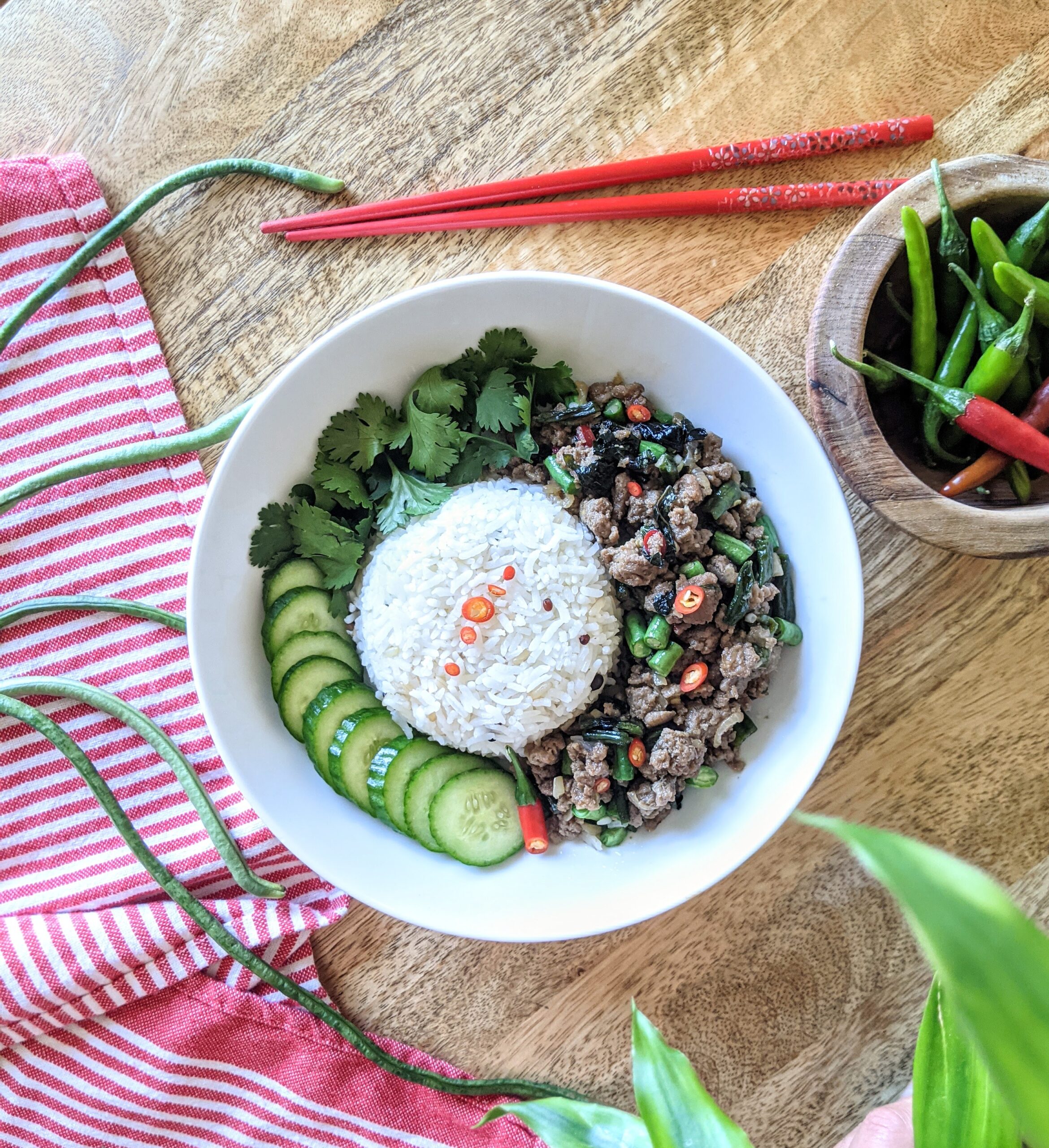 A bowl of spicy Thai basil chicken, steamed Jasmine rice, sliced cucumbers, and fresh cilantro; garnished with sliced Thai chili and fermented black garlic. Long beans and a small bowl of Thai chilies are also pictured.