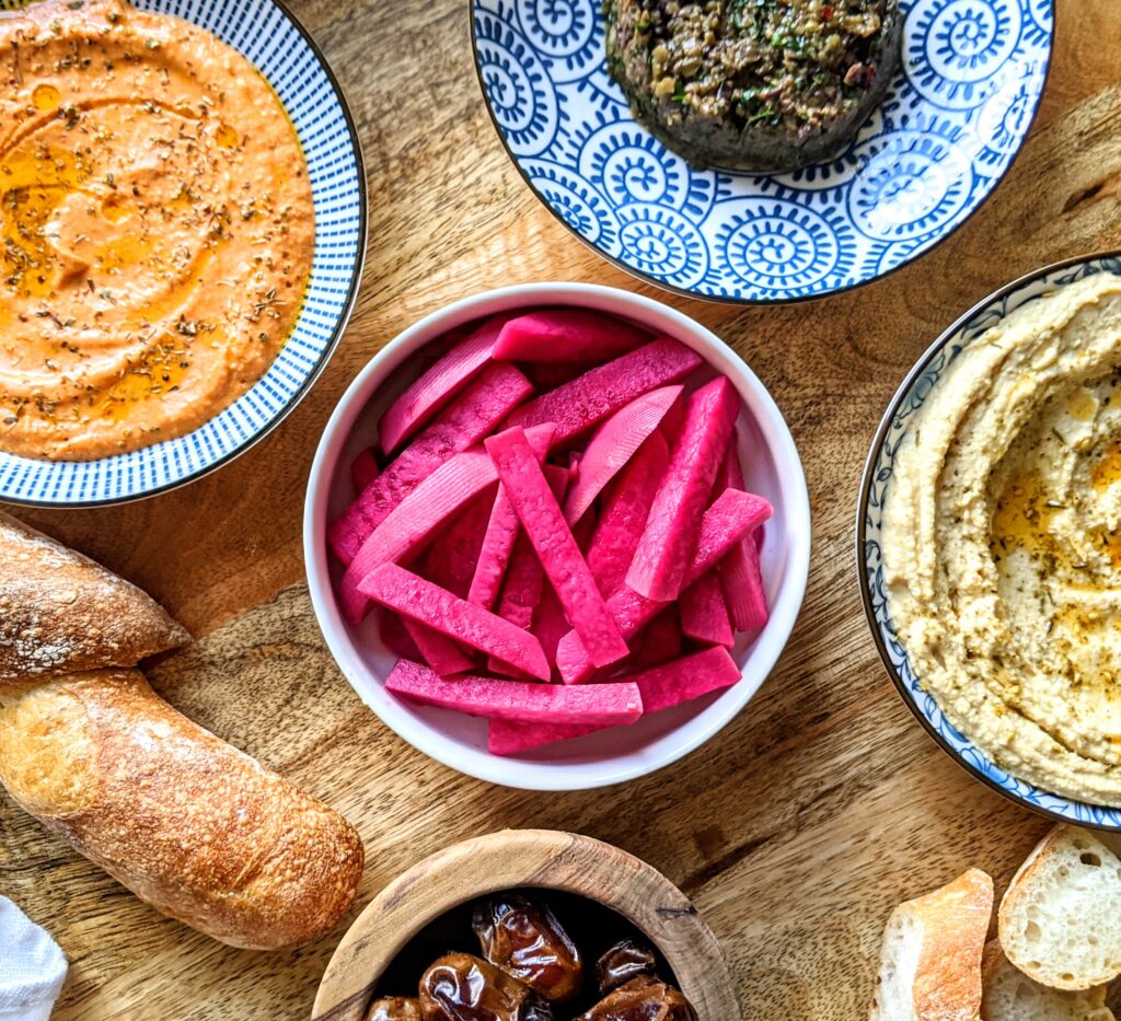 Vibrant pink sticks of Middle eastern pickled turnips. Served alongside other mezes like hummus, olive tapenade, spicy roasted red pepper and feta dip, dates, and a baguette.