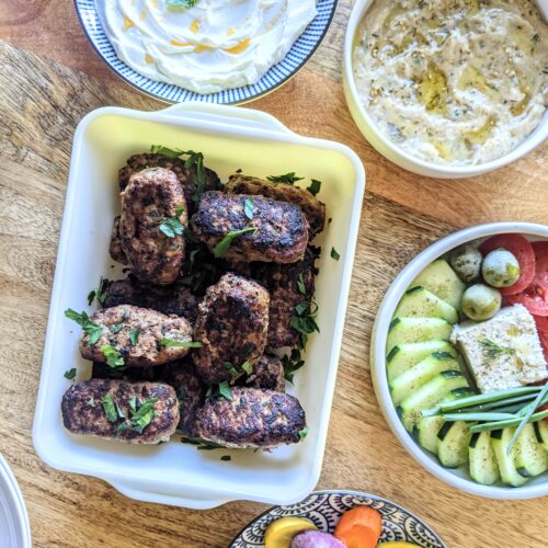 A casserole dish full of log shaped Albanian beef Qofte Meatballs. Served alongside labneh, hummus, a village salad, and pickled vegetables.