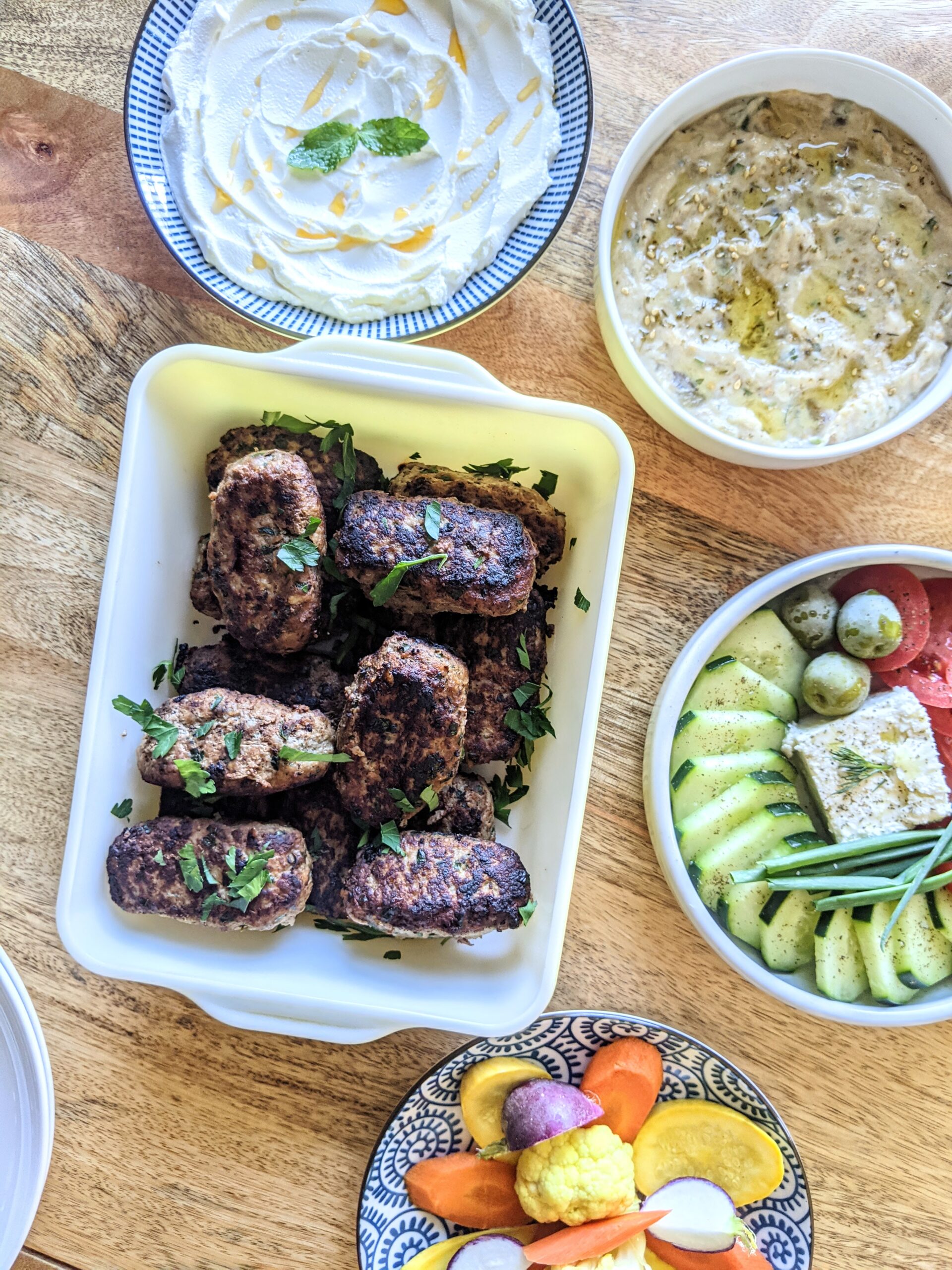 A casserole dish full of log shaped Albanian beef Qofte Meatballs. Served alongside labneh, hummus, a village salad, and pickled vegetables.