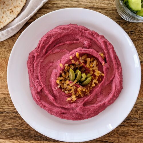 Vibrant pink beetroot hummus swooshed and swirled onto a white plate; garnished with chopped preserve lemon and sliced Castelvetrano olives. Served alongside warm pita bread and Persian cucumber spears.