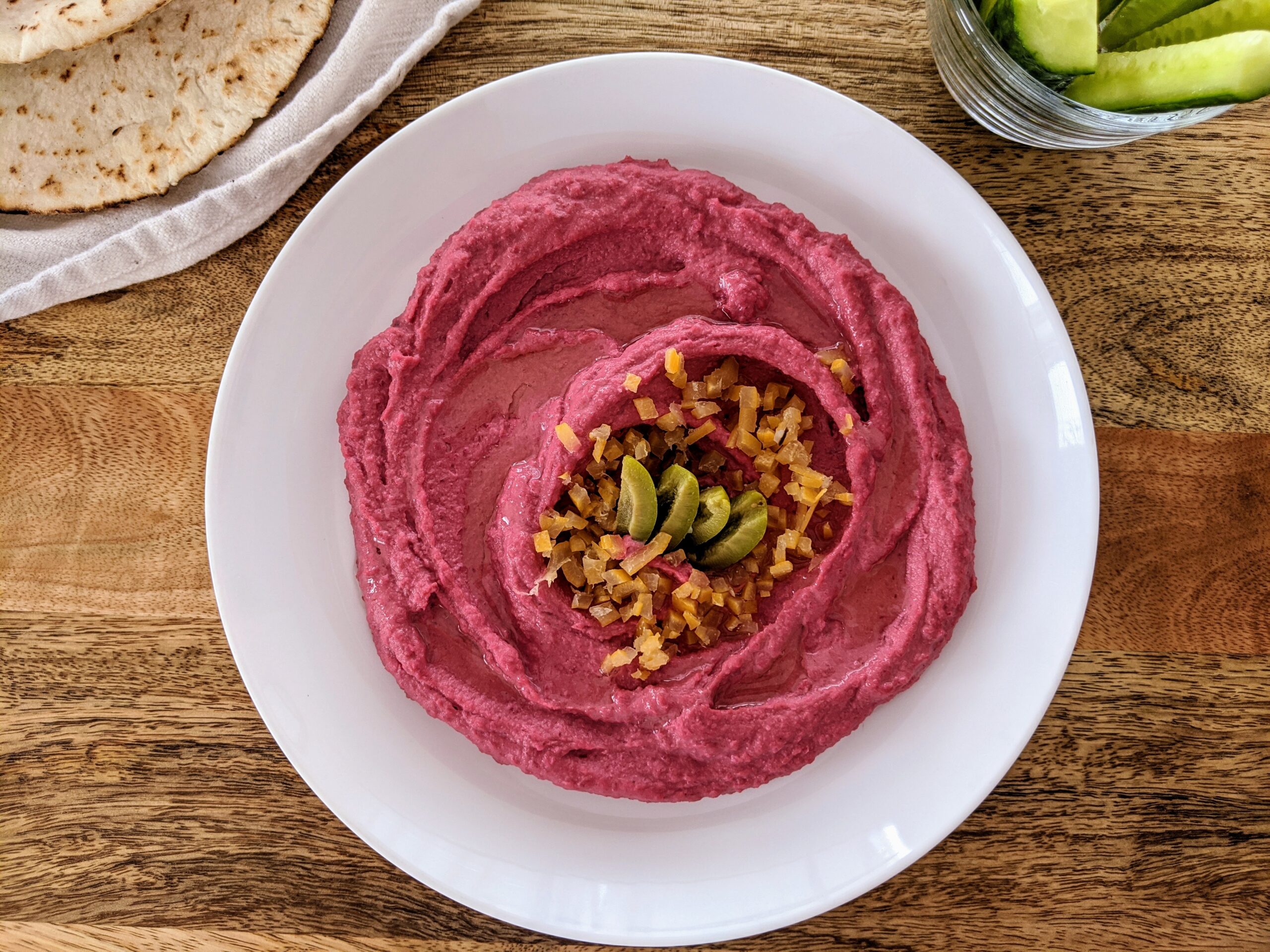 Vibrant pink beetroot hummus swooshed and swirled onto a white plate; garnished with chopped preserve lemon and sliced Castelvetrano olives. Served alongside warm pita bread and Persian cucumber spears.
