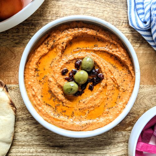 A shallow bowl full of orange roasted red pepper and sun-dried tomato hummus. Garnished with chopped sun-dried tomatoes, 3 Castelvetrano olives, and dried Greek oregano.