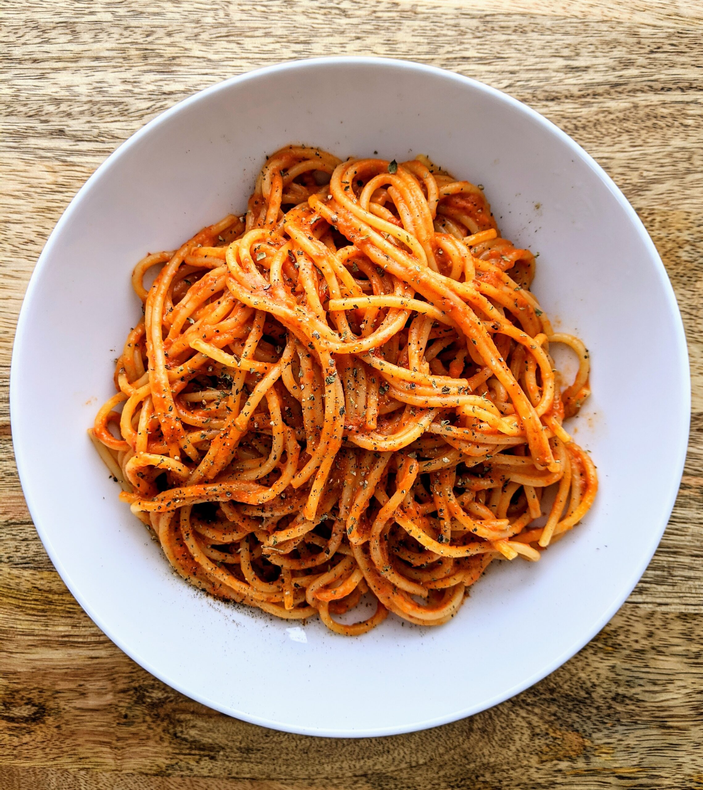 A bowl of spaghetti coated in a bright red roasted red pepper and tomato sauce. Garnished with a dusting of dried oregano.
