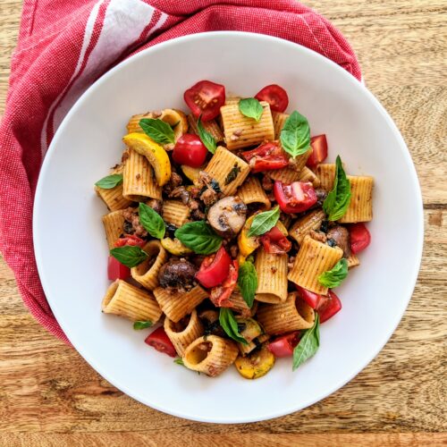 A vibrant bowl of pasta laced with yellow zucchini, mushrooms, tomatoes, and savory sausage. Garnished with fresh basil leaves.