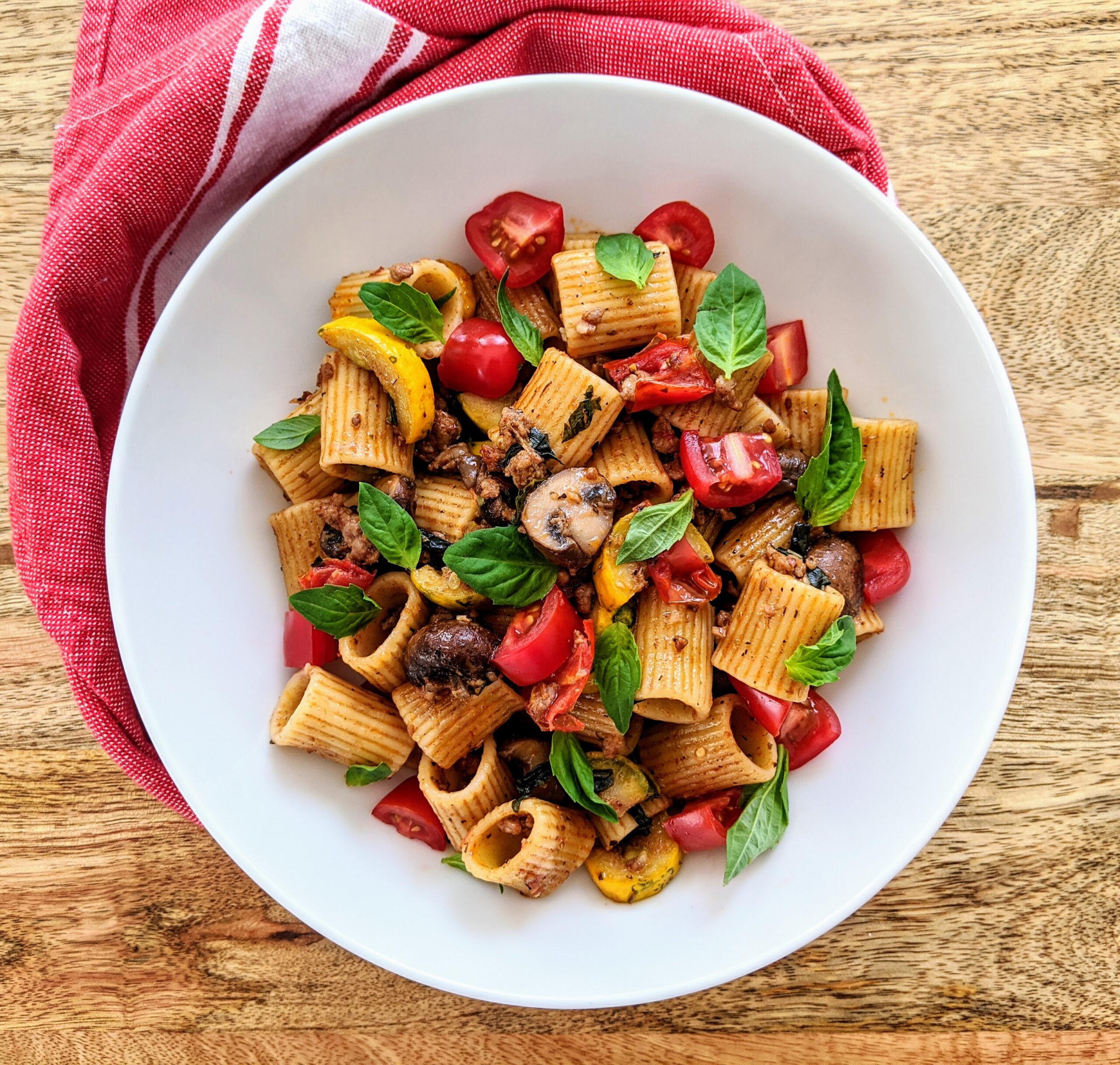 A vibrant bowl of pasta laced with yellow zucchini, mushrooms, tomatoes, and savory sausage. Garnished with fresh basil leaves.