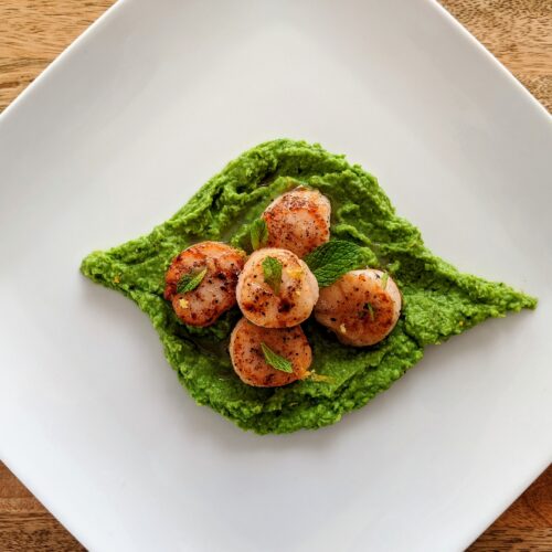 A mint leaf shape of pea purée, topped with five seared scallops, served on a contrasting white square plate; garnished with small mint leaves.
