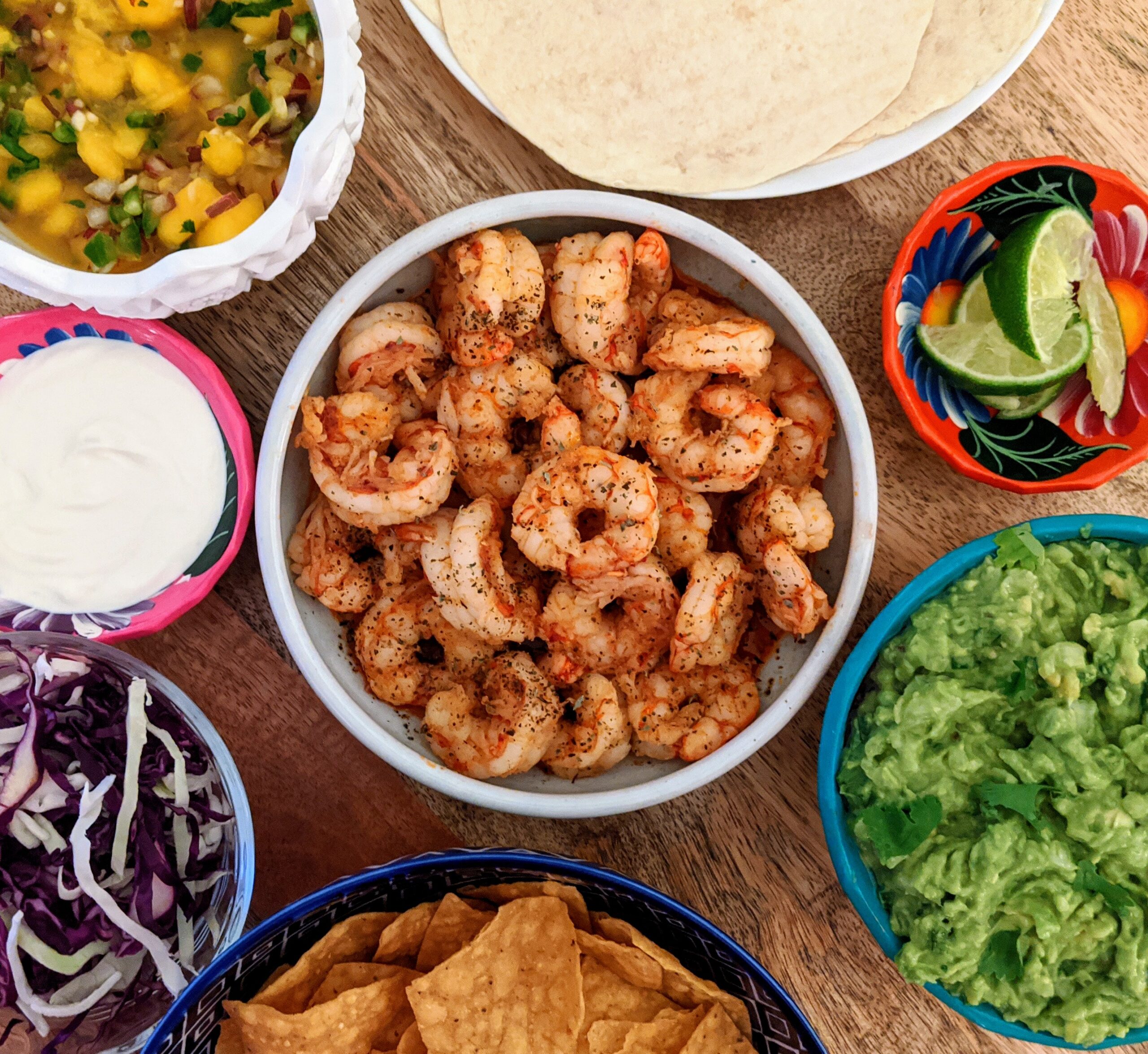 A bowl of plump and spicy shrimp. Surrounded by all of the things you would need to make tacos, like guacamole, pineapple salsa, lime crema, warm tortillas, shredded cabbage, and lime wedges.
