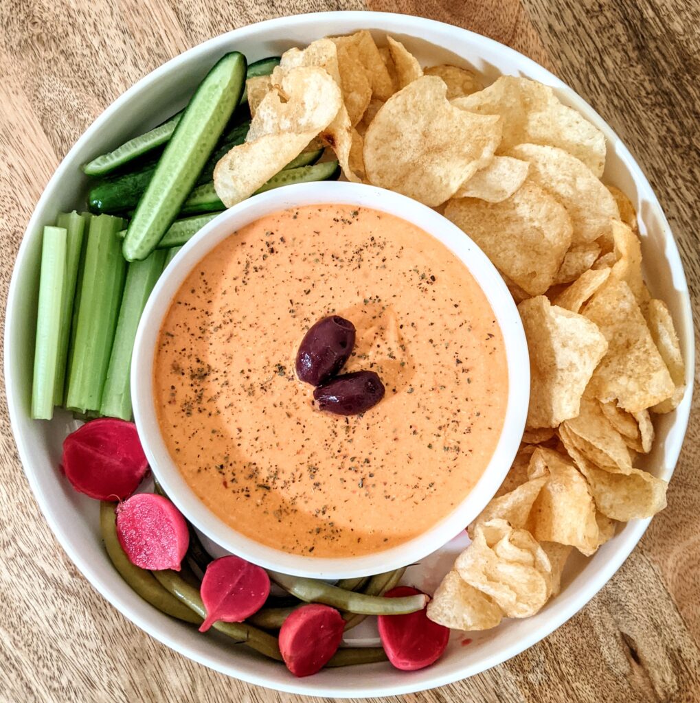 A plate of spicy roasted red pepper and feta dip (tirokafteri) surrounded by kettle cooked potato chips, pickled radishes, and raw celery sticks and Persian cucumber spears. The dip is garnished with whole Kalamata olives and a dusting of dried Greek oregano.