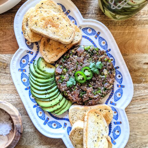 Tuna Tartare plated in a circular ring mold. Garnished with two slices of jalapeño and fresh cilantro. Served alongside thinly sliced avocado and fermented black garlic toasts.