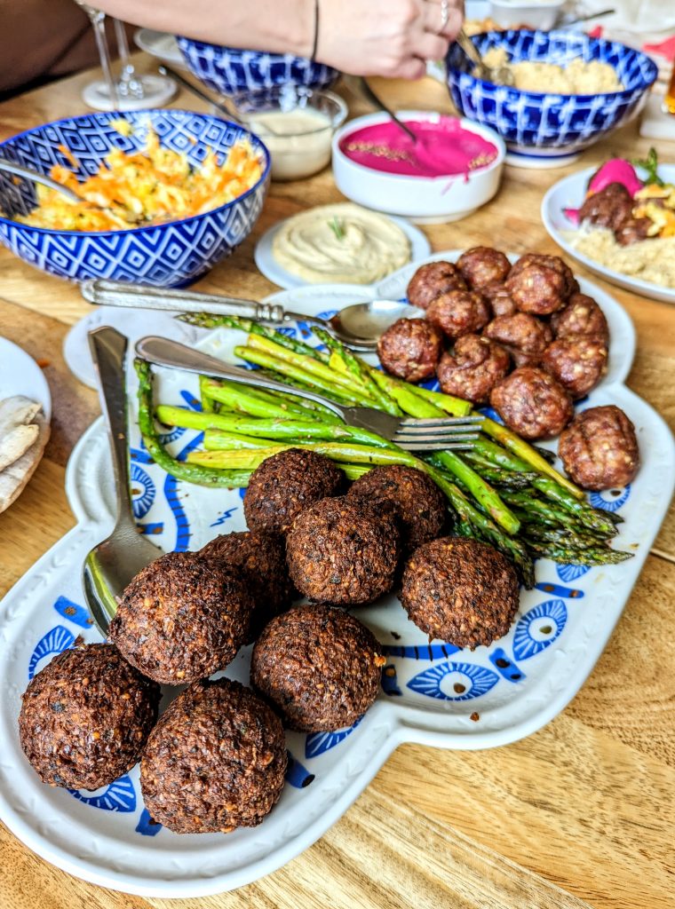 A table full of food including falafel, meatballs, roasted asparagus, a variety or salads and dips. 