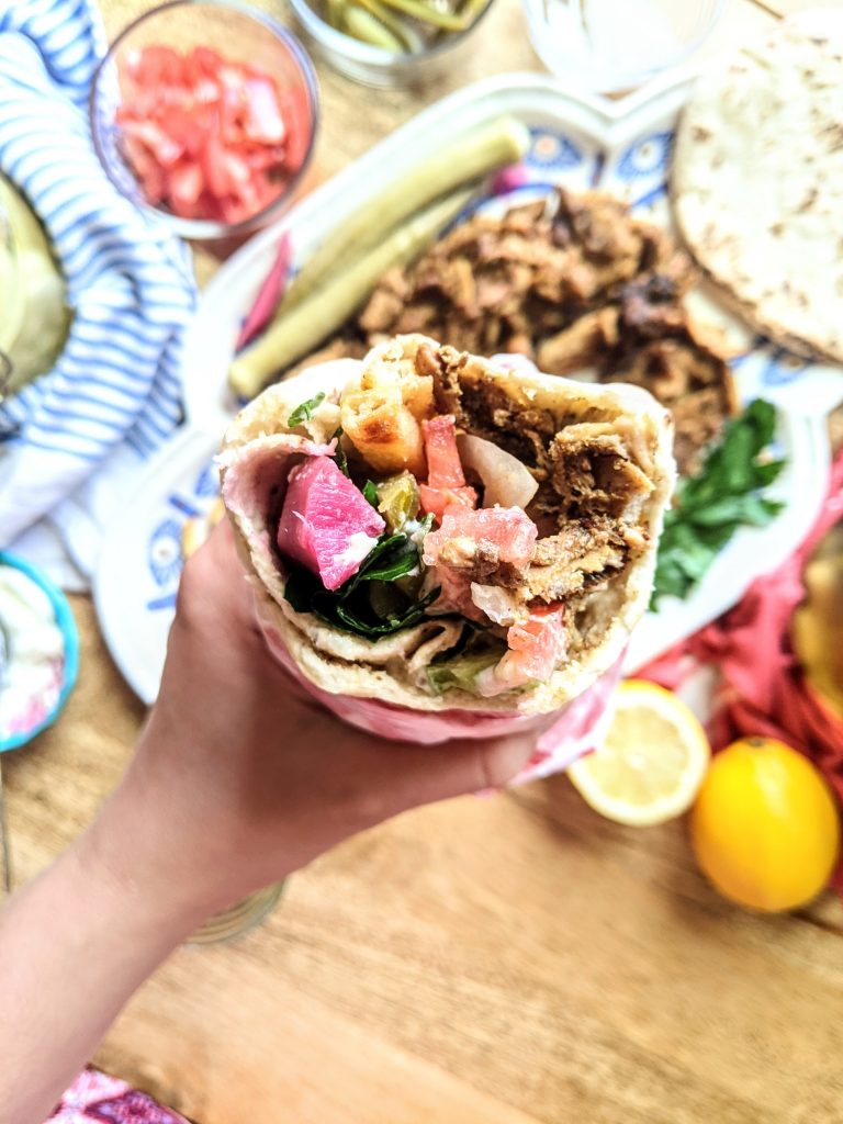 A person holding a chicken shawarma wrap overtop of a shawarma platter.