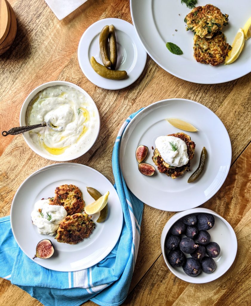 A flat lay with three plates full of Zucchini and Feta Fritters, Turkish pickles, yogurt sauce, lemon wedges, and fresh Mission figs.
