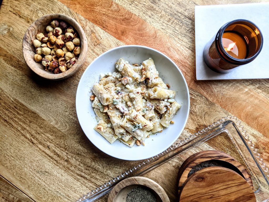 An aerial landscape photo with a bowl of blue cheese and hazelnut rigatoni, a small wooden bowl of whole hazelnuts, and a light candle jar.