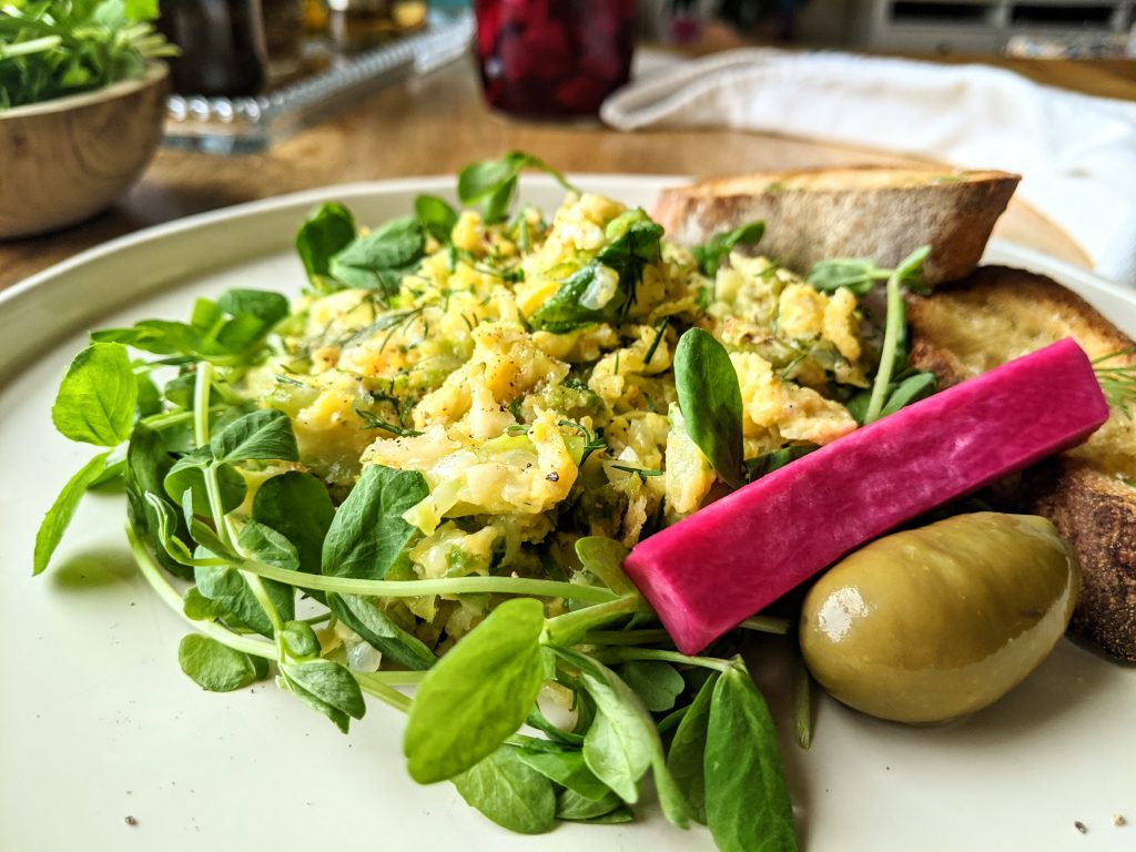 Close up photo of a Brussels Sprouts Scramble with pea shoots, pickled turnips, olives, and sourdough toasts.